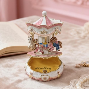 Può includere: Un carillon a carosello decorativo in rosa, bianco e oro. La giostra presenta cavalli in miniatura e un tetto a strisce. Il nome "Hadley" è inciso sulla base. Il carillon è su una superficie rosa.