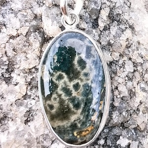 May include: Oval pendant with a silver-colored bail and setting. The pendant features a polished, speckled gemstone in shades of green, gray, and brown. The stone has a glossy finish and is set against a textured background.