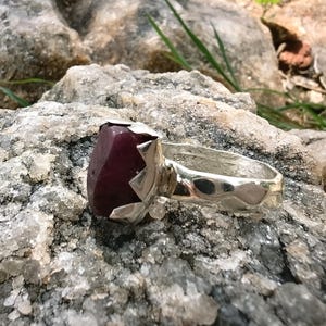 May include: A silver ring featuring a large, faceted, deep red gemstone. The ring's setting has a unique, angular design, and the band has a textured surface. The ring is displayed on a rough, gray rock surface.