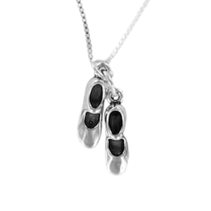 May include: A silver necklace featuring a charm of two ballet slippers. The slippers have black interiors and are connected by a loop to a delicate chain. The pendant is a charming accessory for dance enthusiasts.