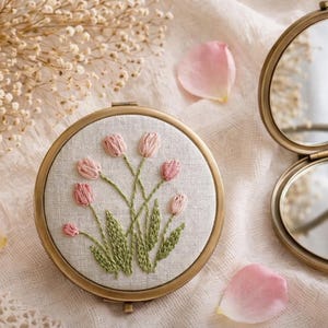 Op de afbeelding: Een ronde compacte spiegel in vintage-stijl met een messing afwerking. De compact heeft een geborduurd bloemmotief van roze tulpen op een crèmekleurige achtergrond. De spiegel is open en onthult twee reflecterende oppervlakken.