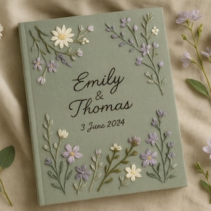 Può includere: Libro degli ospiti di nozze verde salvia con dettagli floreali ricamati. La copertina presenta i nomi "Emily & Thomas" e la data "3 giugno 2024". Fiori e verde decorativi circondano il libro, con ulteriori accenti floreali.