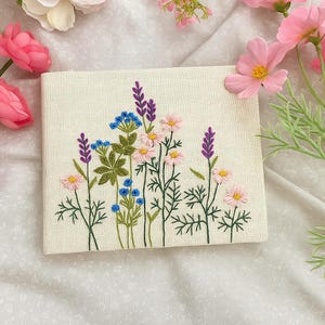 Pode incluir: Um quadrado bordado creme com um design floral. O bordado apresenta flores rosa, roxas e azuis com caules e folhas verdes. As flores estão dispostas em um padrão naturalista, criando uma estética delicada e artesanal.