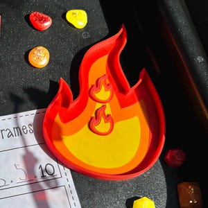 May include: A red and yellow flame-shaped cookie cutter with a smaller flame cutout. Several colorful, glittery resin pieces are scattered around the cutter. A pencil and a worksheet with math problems are also visible.