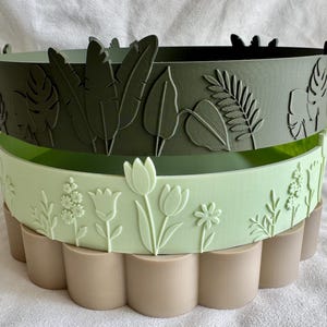 May include: A stack of three cylindrical containers with distinct exterior textures. The top container is dark olive green with tropical leaf reliefs. The middle container is light mint green with floral motifs. The bottom container is beige with a scalloped base.