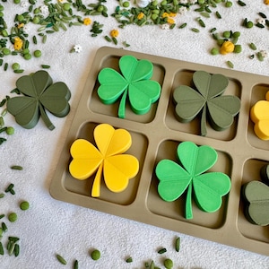 May include: A tan rectangular tray with square compartments containing three-dimensional shamrock shapes in bright green, olive green, and yellow. One additional dark green shamrock sits outside the tray, surrounded by scattered green and yellow bits.