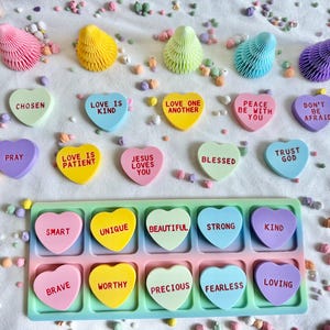 May include: An assortment of colorful heart-shaped candies with inspirational messages like "Chosen," "Love is Kind," and "Trust God." The candies are in various colors, including pink, yellow, blue, and purple, and are arranged on a white surface.
