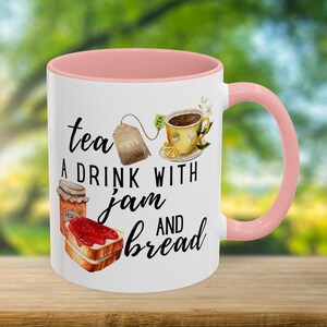 Puede incluir: Taza de cerámica blanca con interior y asa rosas. La taza presenta el texto "tea A DRINK WITH jam AND bread" con ilustraciones de té, mermelada y pan. El fondo es verde borroso.