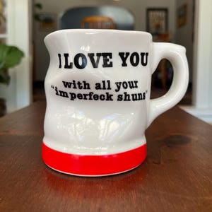 May include: A white ceramic mug with a handle and a red band at the base. The mug has a slightly indented, dimpled texture. Black text printed on the front reads: I LOVE YOU with all your "imperfeck shuns". The mug has a glossy, smooth finish.