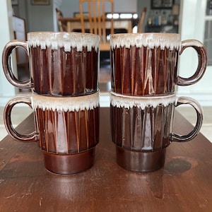 May include: Four stacked brown ceramic mugs with a ridged design and a white, textured rim. The mugs have large, curved handles and are arranged on a dark wooden surface. The mugs are a warm brown color.