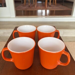 Puede incluir: Cuatro tazas naranjas con interiores blancos dispuestas sobre una superficie de madera. Cada taza tiene un asa redondeada y un acabado liso y brillante. Las tazas están espaciadas uniformemente, mostrando un diseño simple y moderno.