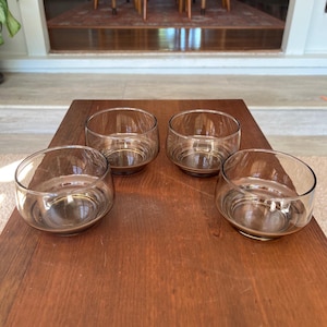 May include: Four clear glass bowls with a rounded shape are arranged on a dark wooden table. The bowls have a simple, modern design, and the glass appears to be slightly tinted. The bowls are empty and ready for use.