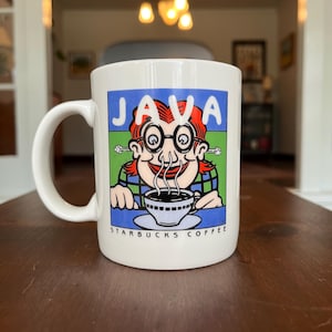 May include: White ceramic coffee mug with a cartoon illustration of a person with red hair and glasses, steam rising from a coffee cup. The mug has the word "JAVA" in blue and the text "STARBUCKS COFFEE".