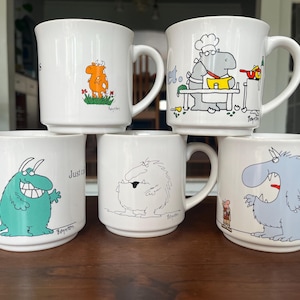 Puede incluir: Una taza de cerámica blanca con un asa negra y el texto "But First, Coffee" en negro, con una fuente de estilo manuscrito. La taza está sobre una superficie blanca.