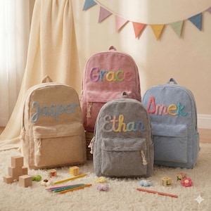 May include: Four corduroy backpacks in beige, pink, grey, and blue, each embroidered with a name in colourful thread. The backpacks have a front pocket and a top handle. Toys and pencils are scattered on the rug.
