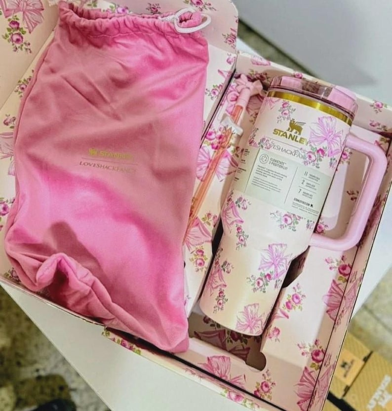 K&ouml;nnte beinhalten: Ein Stanley und LoveShackFancy Kooperationsset bestehend aus einem rosa Samtbeutel mit Kordelzug und einem Edelstahlbecher mit Blumen- und Schleifenmuster. Der Becher enth&auml;lt einen passenden Strohhalm und ein Etikett mit den Stanley und LoveShackFancy Logos.