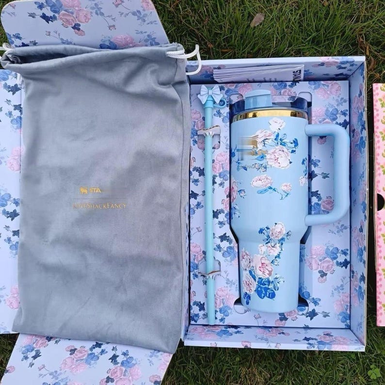 May include: A light blue insulated tumbler with a floral pattern, a matching straw with a bow topper, and a grey drawstring storage bag, all presented in a floral-patterned box. The tumbler features a gold-tone rim and a sturdy side handle.