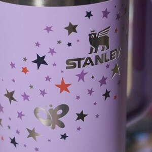 May include: A light purple tumbler featuring a metallic silver Stanley logo with a winged bear icon. The surface is decorated with scattered stars in shades of dark purple, orange, and silver, along with a metallic silver butterfly-shaped graphic.