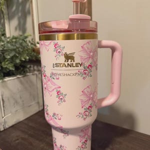 K&ouml;nnte beinhalten: Ein hellrosa Stanley LoveShackFancy Becher mit Blumen- und Schleifenmuster. Der Becher hat einen goldenen Rand, einen rosa Henkel und einen durchsichtigen Strohhalm mit einer rosa Schleife als Aufsatz. Das Stanley-Logo und der LoveShackFancy-Schriftzug sind in Gold gedruckt.