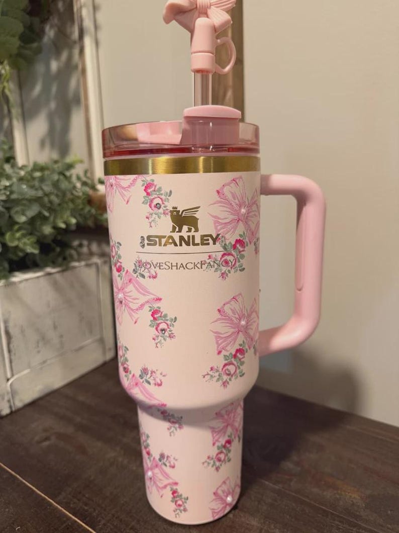 K&ouml;nnte beinhalten: Ein hellrosa Stanley LoveShackFancy Becher mit Blumen- und Schleifenmuster. Der Becher hat einen goldenen Rand, einen rosa Henkel und einen durchsichtigen Strohhalm mit einer rosa Schleife als Aufsatz. Das Stanley-Logo und der LoveShackFancy-Schriftzug sind in Gold gedruckt.
