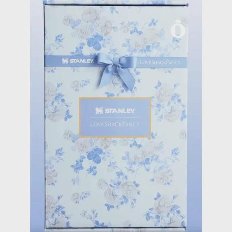May include: A rectangular product box featuring a light blue and white floral pattern. A horizontal blue band across the center displays the text Stanley and LoveShackFancy with a blue ribbon bow. A central label also reads Stanley LoveShackFancy.