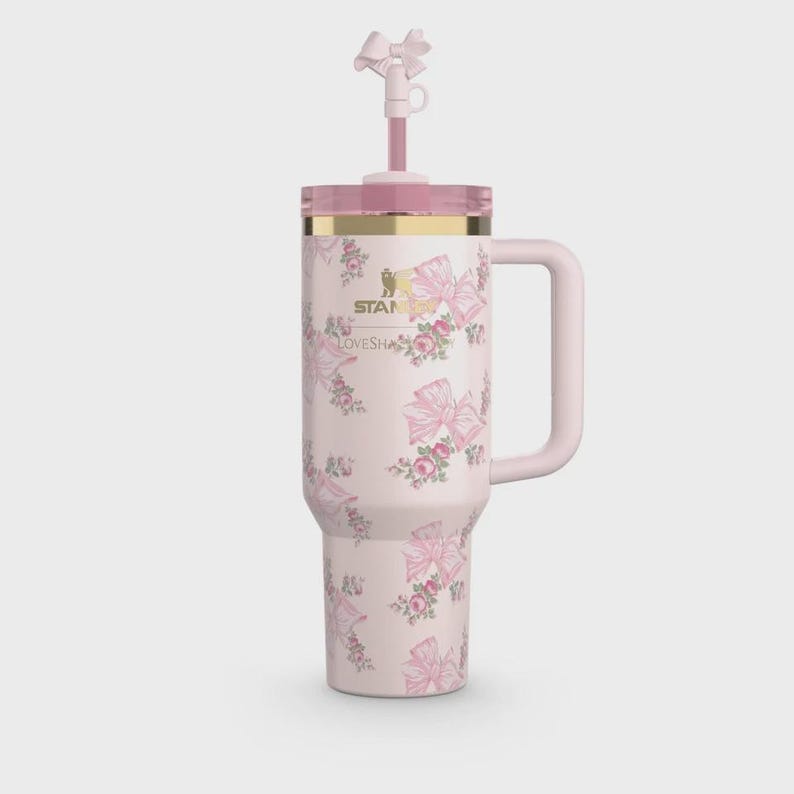 May include: A light pink insulated tumbler with a handle, featuring a repeating pattern of pink bows and small floral clusters. The Stanley logo and LoveShackFancy text are printed in gold. A matching pink straw with a bow topper is included.