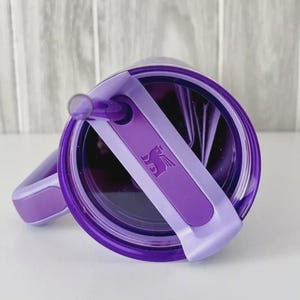 May include: A purple tumbler with a matching lid and straw. The lid features a rotating cover with a debossed winged bear logo. The tumbler has a side handle with a purple grip insert, and the overall design is a deep, translucent purple.