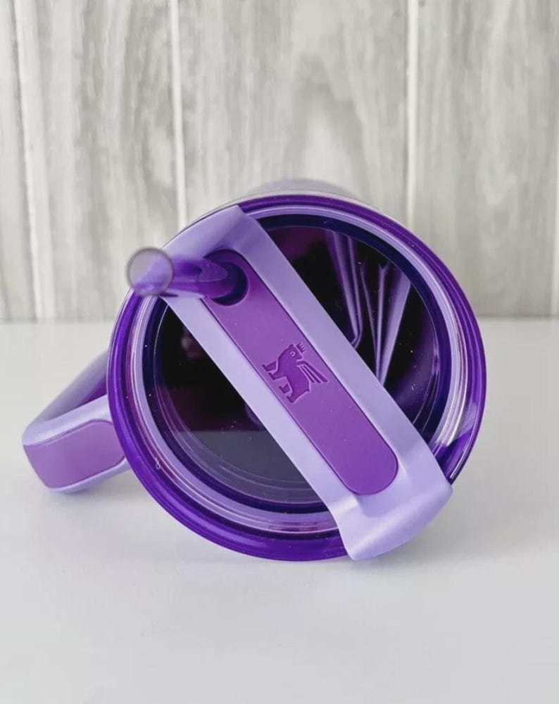 May include: A purple tumbler with a matching lid and straw. The lid features a rotating cover with a debossed winged bear logo. The tumbler has a side handle with a purple grip insert, and the overall design is a deep, translucent purple.
