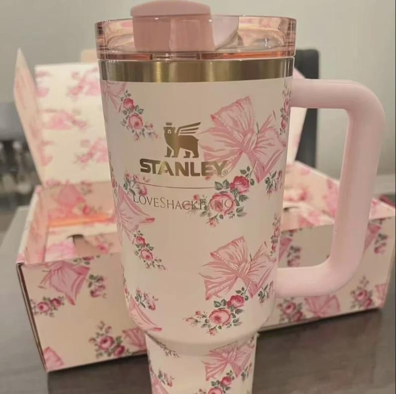 K&ouml;nnte beinhalten: Ein hellrosa Stanley-Becher mit einem LoveShackFancy-Blumen- und Schleifenmuster. Der Becher hat einen goldfarbenen Rand, einen passenden rosa Henkel und einen transparenten Deckel mit rosa Schiebeverschluss. Das Stanley-Logo und der LoveShackFancy-Schriftzug sind in Gold gedruckt.