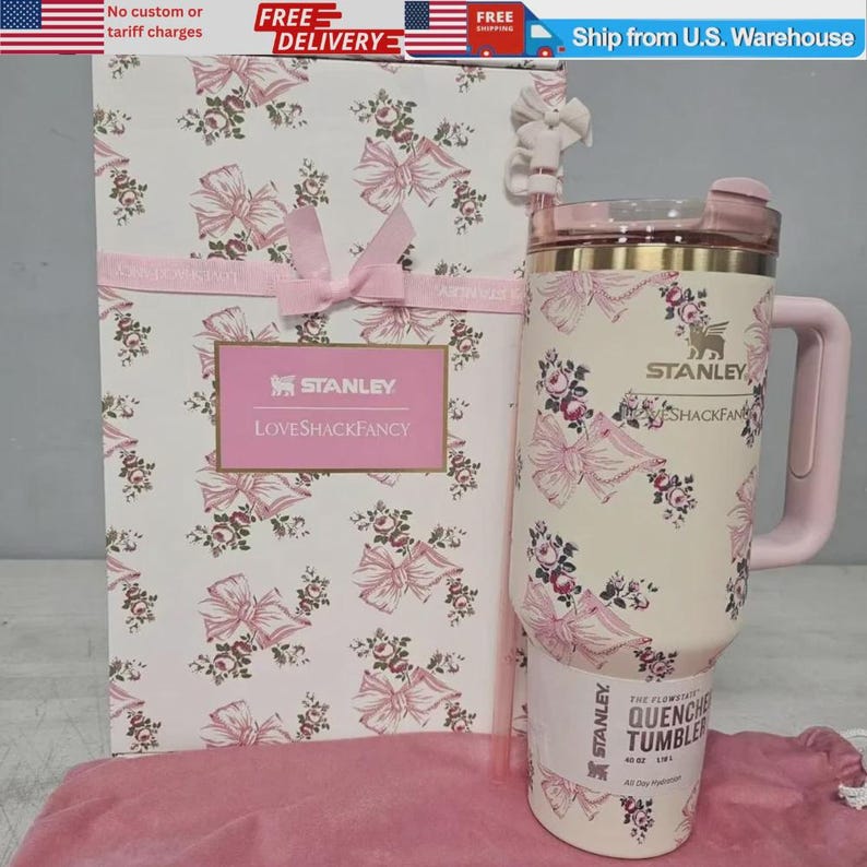 K&ouml;nnte beinhalten: Ein cremefarbener Stanley Quencher Becher mit einem rosa Blumen- und Schleifenmuster, inklusive passendem Henkel, Deckel und Strohhalm. Der Becher steht neben einer hohen, rechteckigen Geschenkbox mit demselben floralen Aufdruck.