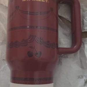 K&ouml;nnte beinhalten: Ein tiefroter Stanley-Becher mit goldenem Rand und passendem rotem Deckel. Der Becher zeigt ein goldenes Logo, ein dekoratives Girlandenmuster und eine Kirschgrafik. Ein wei&szlig;es Etikett am Boden gibt 11 Stunden kalt und 2 Tage eisgek&uuml;hlt an.