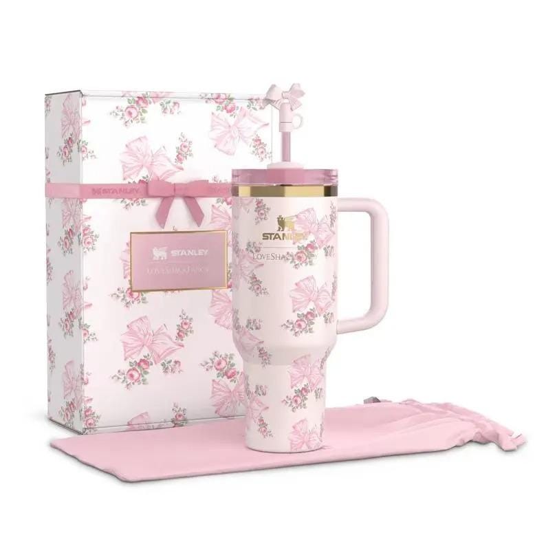 May include: A light pink Stanley tumbler with a floral and bow pattern, a gold rim, a handle, and a matching straw topper. It is displayed next to a rectangular gift box featuring the same pattern and a pink ribbon, plus a soft pink fabric pouch.