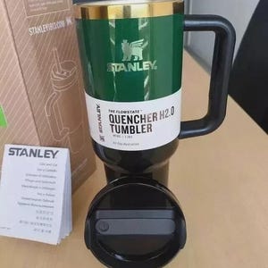May include: A dark green Stanley Flowstate Quencher H2.0 tumbler with a gold rim and black handle. The tumbler features a white label with the text The Flowstate Quencher H2.0 Tumbler 40 oz | 1.18 L. A black lid and a cardboard box are included.