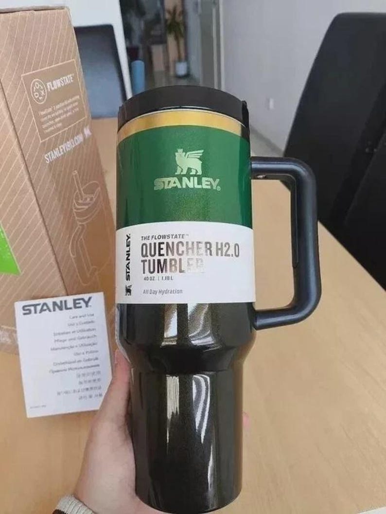May include: A dark green Stanley Quencher H2.0 Flowstate tumbler with a black lid and handle. The tumbler features a white product label with the text Quencher H2.0 Tumbler, 40 oz, and All Day Hydration. A cardboard box and care instruction card are visible.
