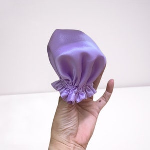 May include: A lilac satin bonnet with a frilled edge. The bonnet is gathered at the base and appears to be made of a soft, flowing fabric. The bonnet is designed to protect hair.