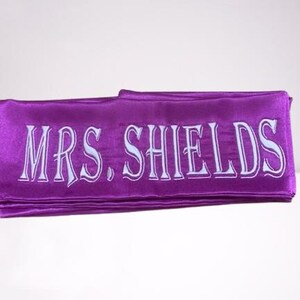 May include: A folded purple satin sash with the text "MRS. SHIELDS" embroidered in white. The sash is a vibrant purple color and appears to be made of a soft, flowing material. The text is in a decorative font.