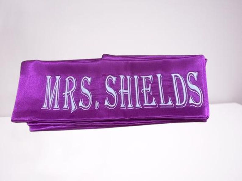 May include: A folded purple satin sash with the text "MRS. SHIELDS" embroidered in white. The sash is a vibrant purple color and appears to be made of a soft, flowing material. The text is in a decorative font.