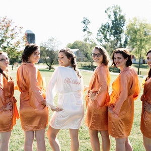 May include: Six people wearing matching peach-colored satin robes pose outdoors on a sunny day. One person wears a white robe with "Mrs. Steer" embroidered on the back. The background includes green grass, trees, and a barn.