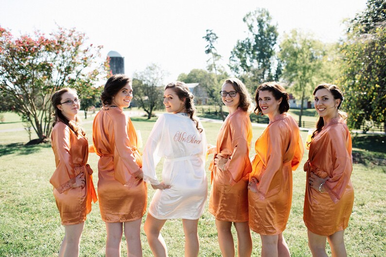 May include: Six people wearing matching peach-colored satin robes pose outdoors on a sunny day. One person wears a white robe with "Mrs. Steer" embroidered on the back. The background includes green grass, trees, and a barn.