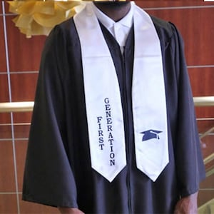 May include: A black graduation gown with a white sash. The sash features the words "FIRST GENERATION" in blue text and a blue graduation cap icon. The gown is worn over a white collared shirt.