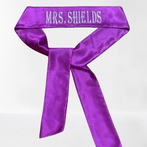 May include: A vibrant purple satin head wrap with the embroidered text "MRS. SHIELDS" in white. The head wrap is tied in a bow shape, with long, flowing tails. The fabric has a slight sheen.