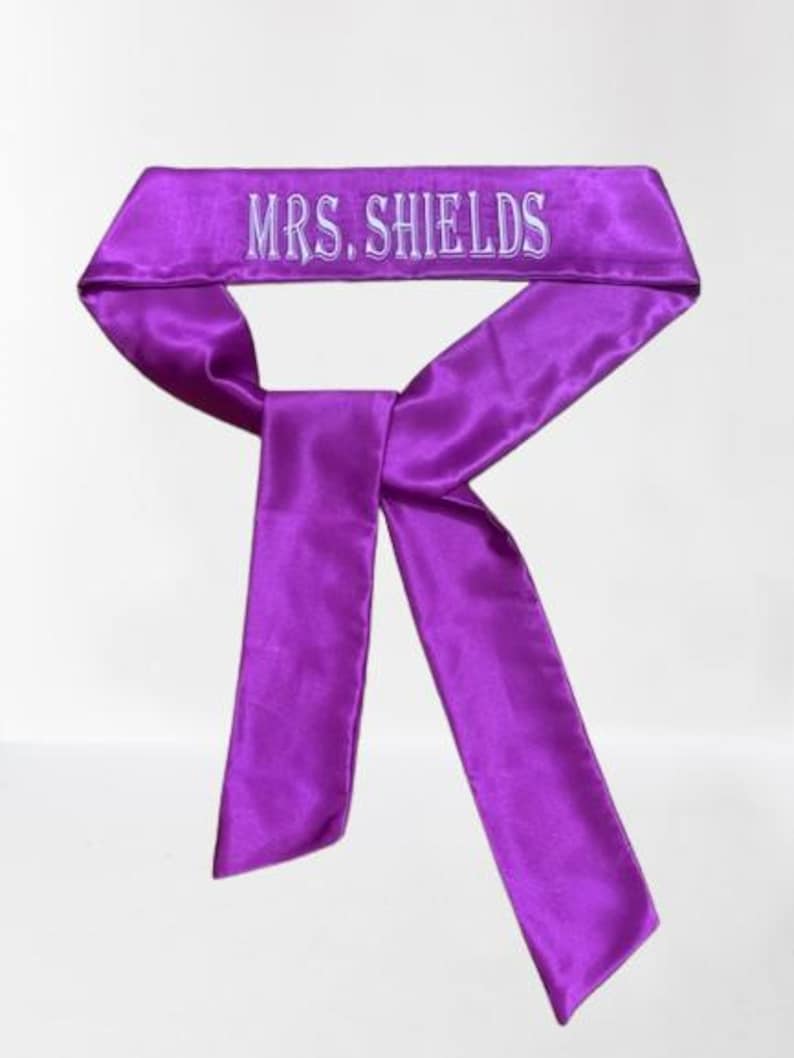 May include: A vibrant purple satin head wrap with the embroidered text "MRS. SHIELDS" in white. The head wrap is tied in a bow shape, with long, flowing tails. The fabric has a slight sheen.