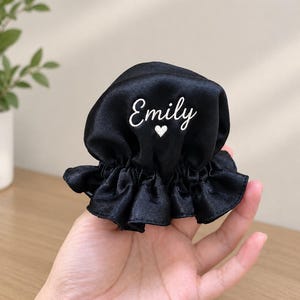Könnte beinhalten: Ein schwarzes Haargummi aus Satin mit gerüschtem Rand. Der Stoff ist mit dem Namen Emily in weißer Schreibschrift bestickt, der sich direkt über einem kleinen, weiß gestickten Herzsymbol in der Mitte des Accessoires befindet.