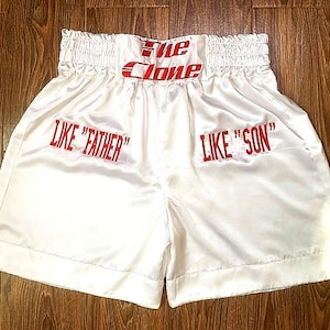 Peut inclure: Short de boxe en satin blanc avec le texte rouge "The Clone" sur la ceinture. Le short porte également les inscriptions "LIKE 'FATHER'" et "LIKE 'SON'" imprimées sur le devant. Le short est présenté sur une surface en bois.