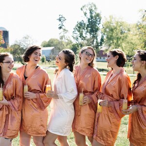 May include: A group of women wearing matching peach-colored satin robes with lace trim, each holding a glass of a yellow beverage. The women are laughing outdoors on a sunny day, suggesting a bridal party or similar event.