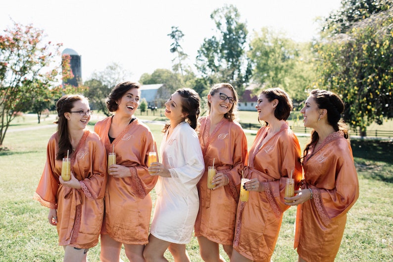 May include: A group of women wearing matching peach-colored satin robes with lace trim, each holding a glass of a yellow beverage. The women are laughing outdoors on a sunny day, suggesting a bridal party or similar event.