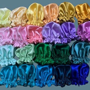 Op de afbeelding: Een collectie van vierentwintig satijnen mutsen gerangschikt in een raster, met een spectrum aan kleuren waaronder geel, perzik, roze, paars, groen, taling en blauw. Elke muts heeft een geplooide elastische rand en een gladde, glanzende afwerking.