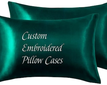 Pode incluir: Duas fronhas de cetim verde esmeralda. As fronhas têm um brilho lustroso e as palavras "Custom Embroidered Pillow Cases" estão escritas em branco na frente.