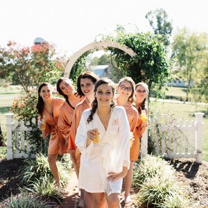 May include: A group of people wearing robes, standing on a brick path. The robes are in shades of peach and white, with lace trim. Each person is holding a drink. The setting is outdoors, with a white picket fence and greenery.