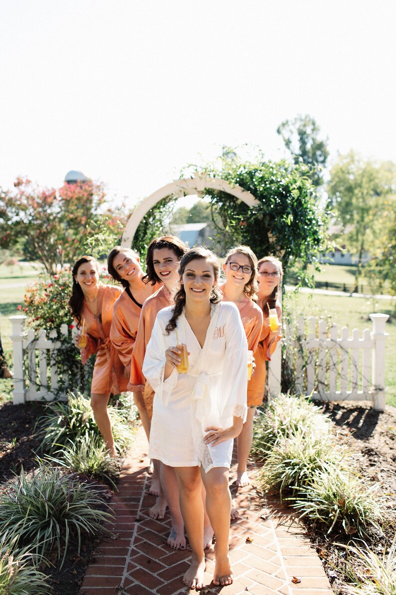 May include: A group of people wearing robes, standing on a brick path. The robes are in shades of peach and white, with lace trim. Each person is holding a drink. The setting is outdoors, with a white picket fence and greenery.